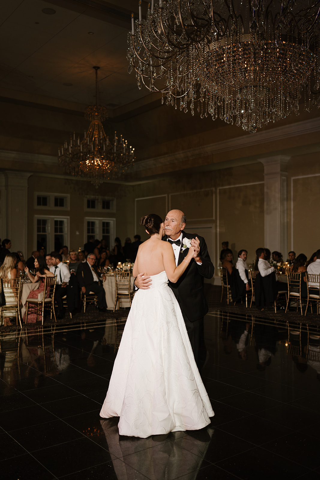 Father kissing his daughter's cheek during an emotional father-daughter dance under crystal chandeliers.